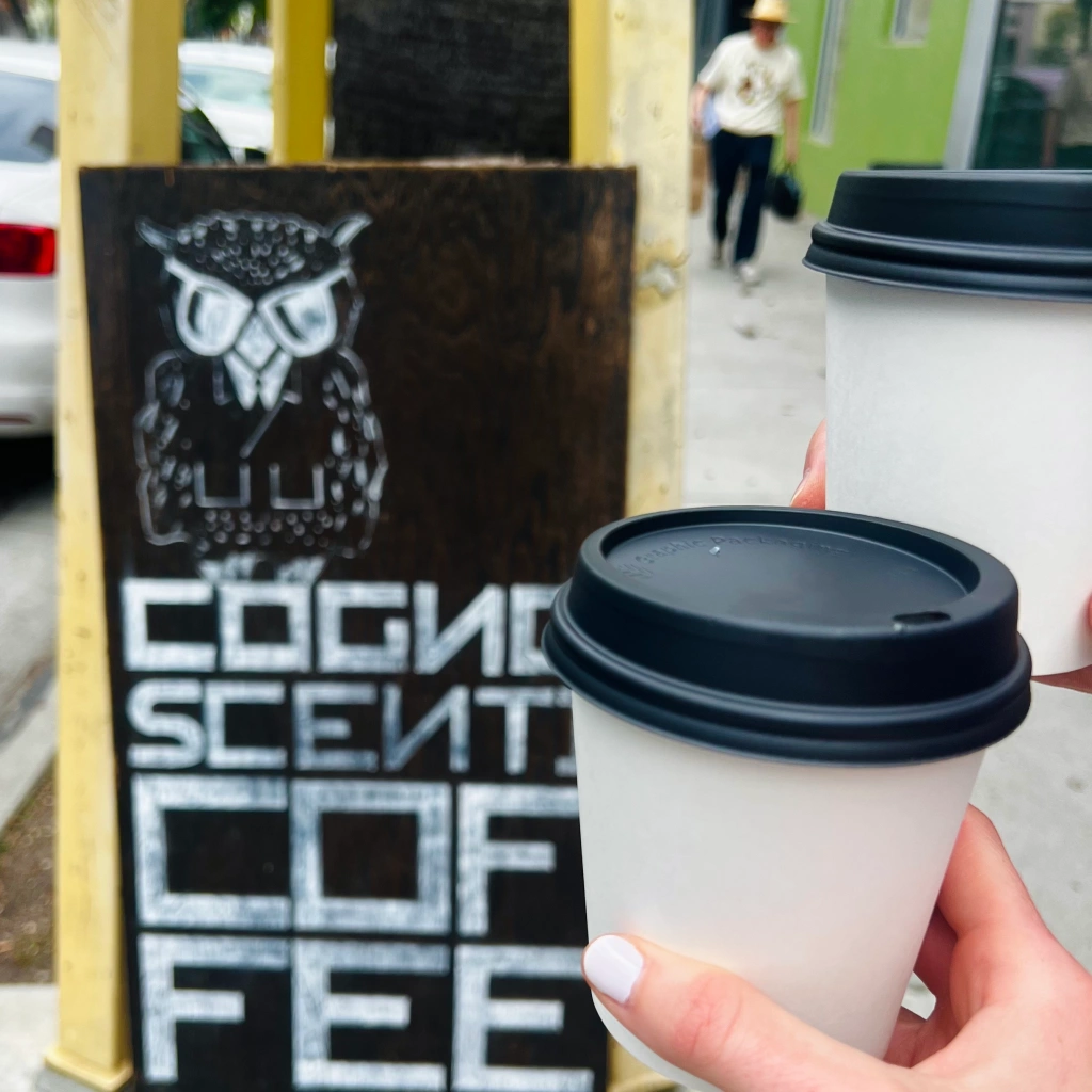 Hand with white nail polish holding hot coffee cup in front of black and white A-frame that reads COGNO SCENTI COF FEE with a painted owl perched on top. Man wearing beige fedora hat, white t-shirt, and black jeans walking in the background.