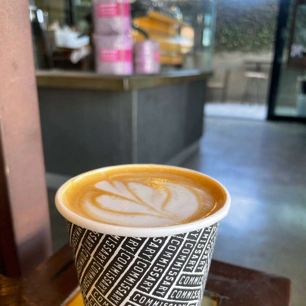 Latte art on top of hot oat latte beverage in a disposable cup inside of Coffee Commissary with three stacked 5-pound bags of coffee on the counter next to the espresso machine in the background, with a view of the patio in the far back blurred out 