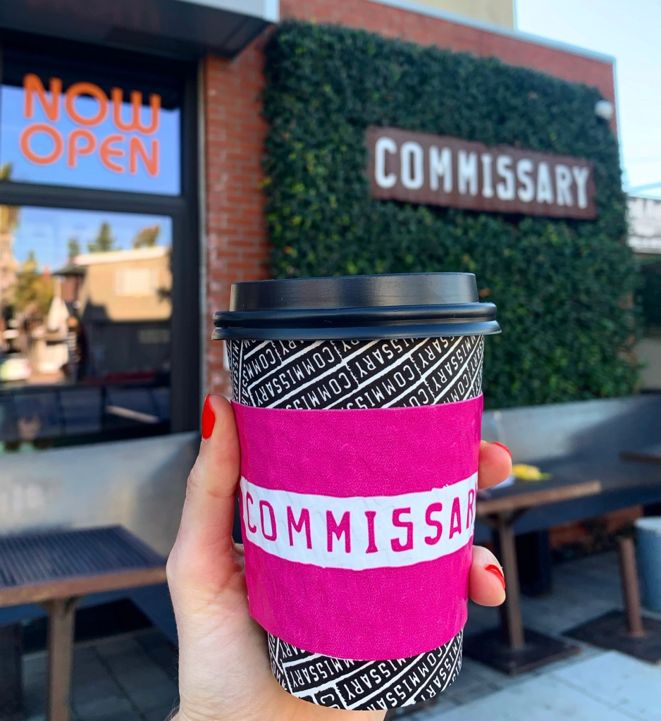Hand holding closed disposable cup with a lid and hot pink sleeve outside of the Coffee Commissary sign on Motor Ave