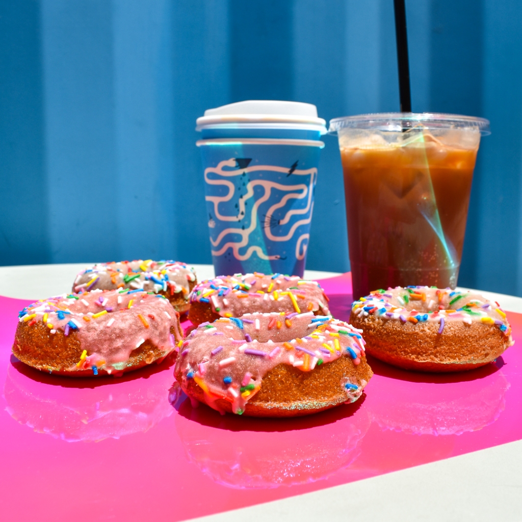 Five pink donuts with sprinkles on a hot pink paper on a table in front of two different coffee beverages, an iced espresso tonic on the right and a hot latte in a hot blue cup with a maze on the left. In front of a bright blue shipping container at Play Coffee.