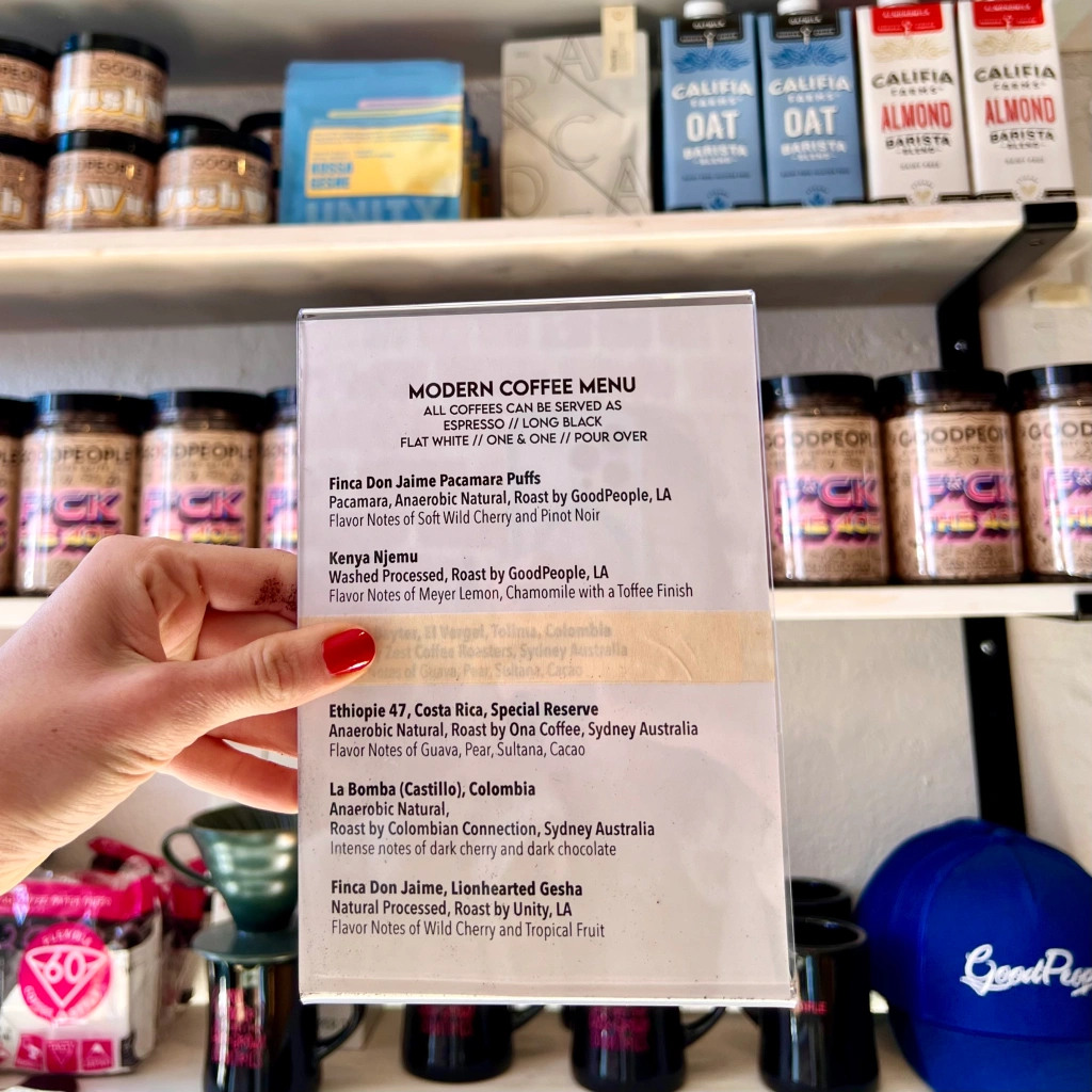 Modern Coffee Menu printed sign available at GoodPeople on Santa Monica Blvd. Containing the list of coffees that can be served as espresso, flat white, or pour over, including the roaster, region, processing, and flavor notes. Sign held in front of retail shelf containing oat milk, almond milk, and bags of coffee.