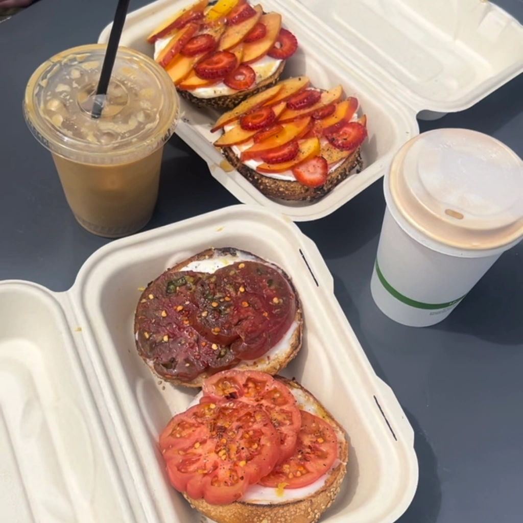The Scarlett bagel with heirloom tomato, lemon zest, chili flakes and vegan cream cheese on a gluten free bagel, a hot latte, an iced latte, and the Pre-Jam with strawberry, nectarine, cream cheese, and honey on a bagel on a table at Layla Bagels in Santa Monica