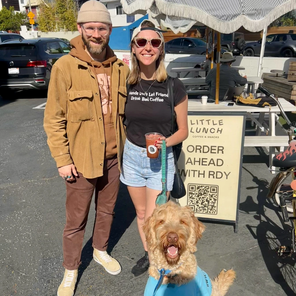 Meagan McCall and Chris McColl standing in the Little Lunch parking lot in front of their sign, picnic tables, and umbrella with the smiling Labradoodle Olaf 