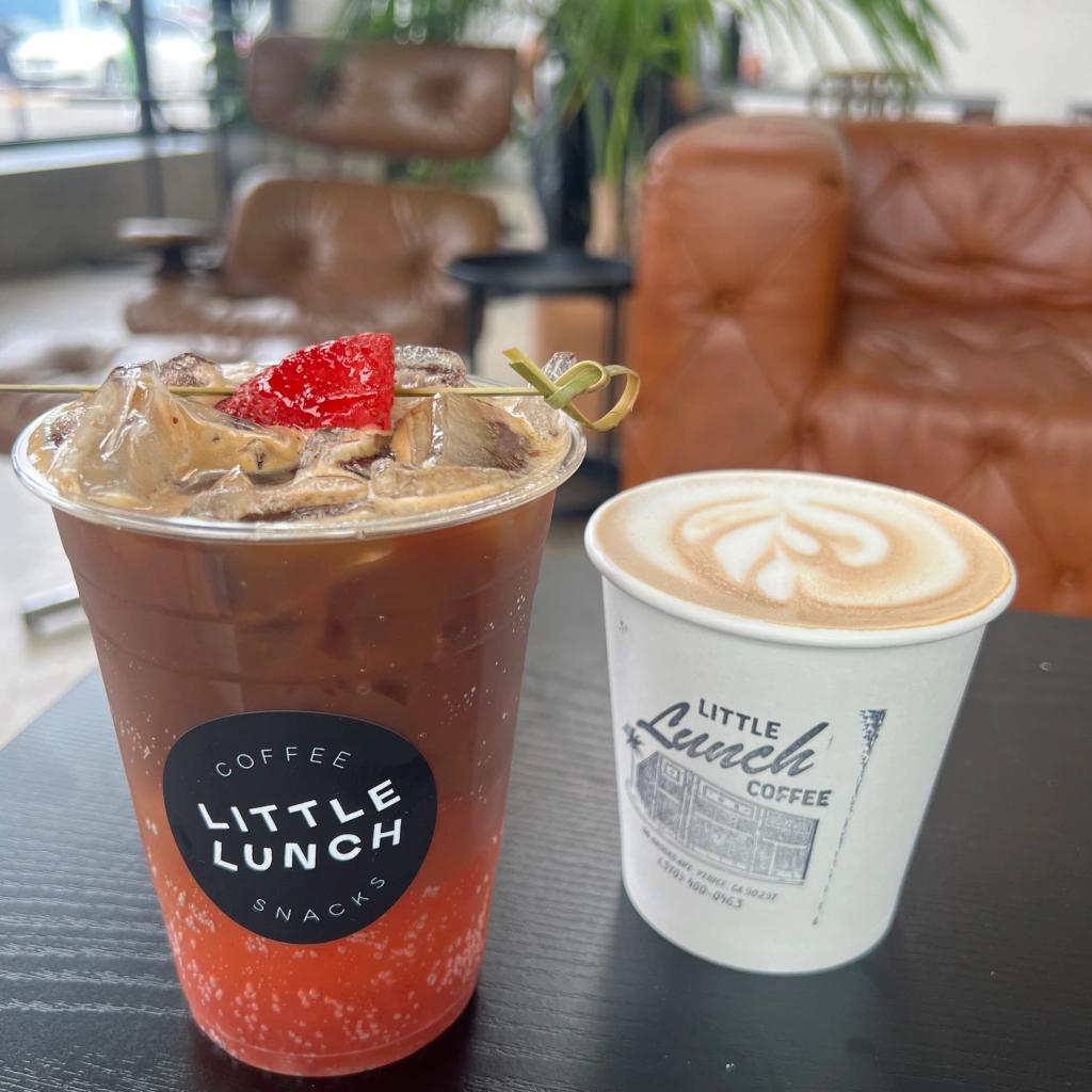 Specialty strawberry beverage and GoodMylk beverage on a table in the Little Lunch indoor seating area in front of a brown leather couch and a brown leather chair 