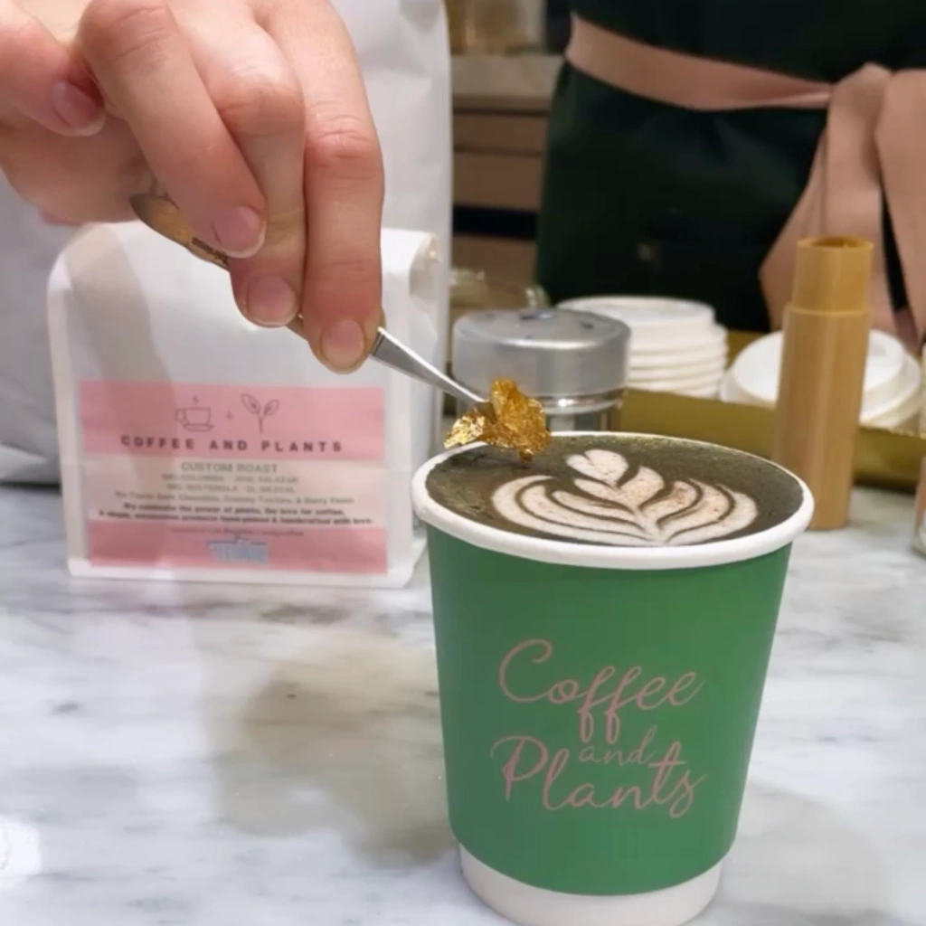 Gold leaf being placed with tweezers on top of a hot 24k charcoal latte in a green cup that reads Coffee and Plants in front of a pink and white retail coffee bag 