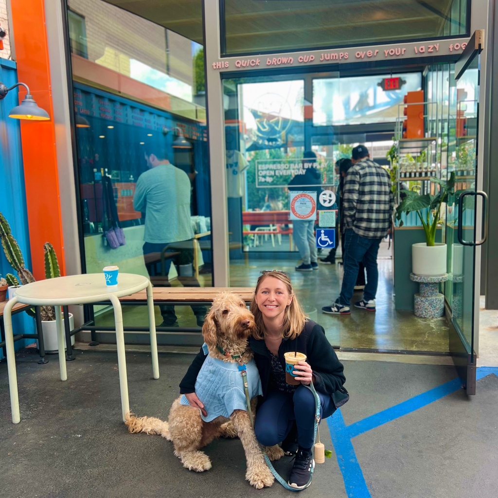 Smiling blonde white girl in black North Face jacket, navy leggings, and black and white Adidas shoes holding apricot-colored labradoodle dog in a light blue knit sweater in front of Play Coffee glass walls. Girl is holding an iced espresso tonic beverage in her hand with the leash draped on her wrist.