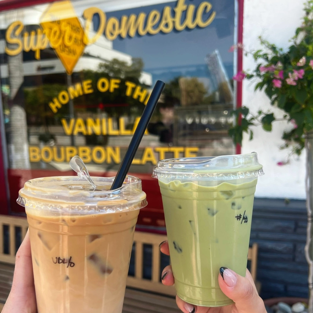 Two hands cheersing with two different beverages, the left beverage being an iced vanilla bourbon latte with Ghost Town oat milk, and the right green beverage being an iced matcha latte. Hands in front of the Home of the Vanilla Bourbon Latte sign in the window of Super Domestic Coffee
