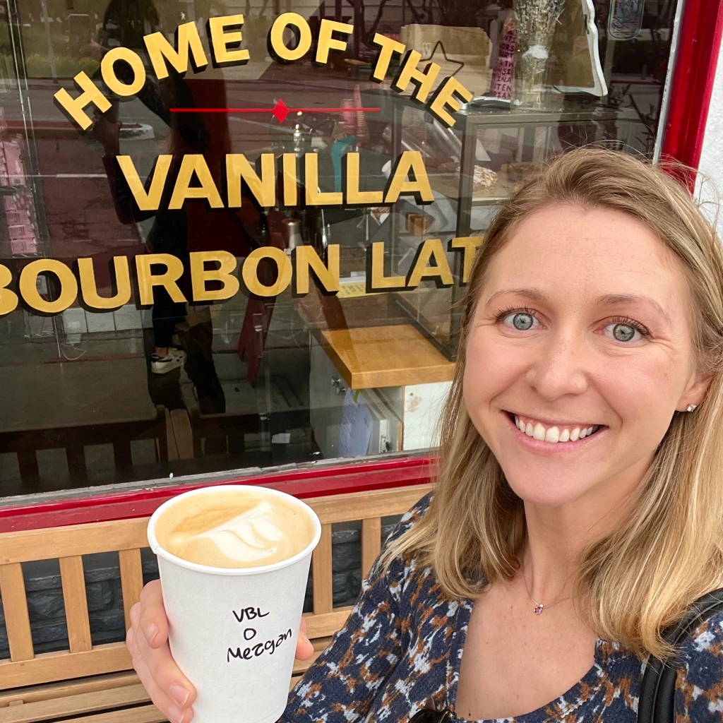 Smiling blonde white girl holding a vanilla bourbon latte with oat milk in front of the Home of the Vanilla Bourbon Latte sign in the window of Super Domestic Coffee