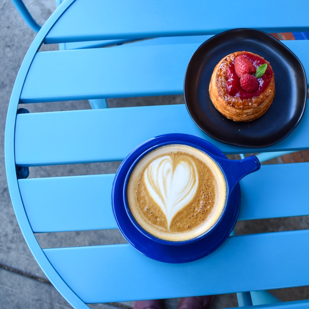 Bright blue metal table containing a hot latte in a for-here cobalt blue cup with a cobalt blue saucer, with a heart shape in the latte art. A berry dessert on a plate on the table at Syndicate Coffee + Bakery.